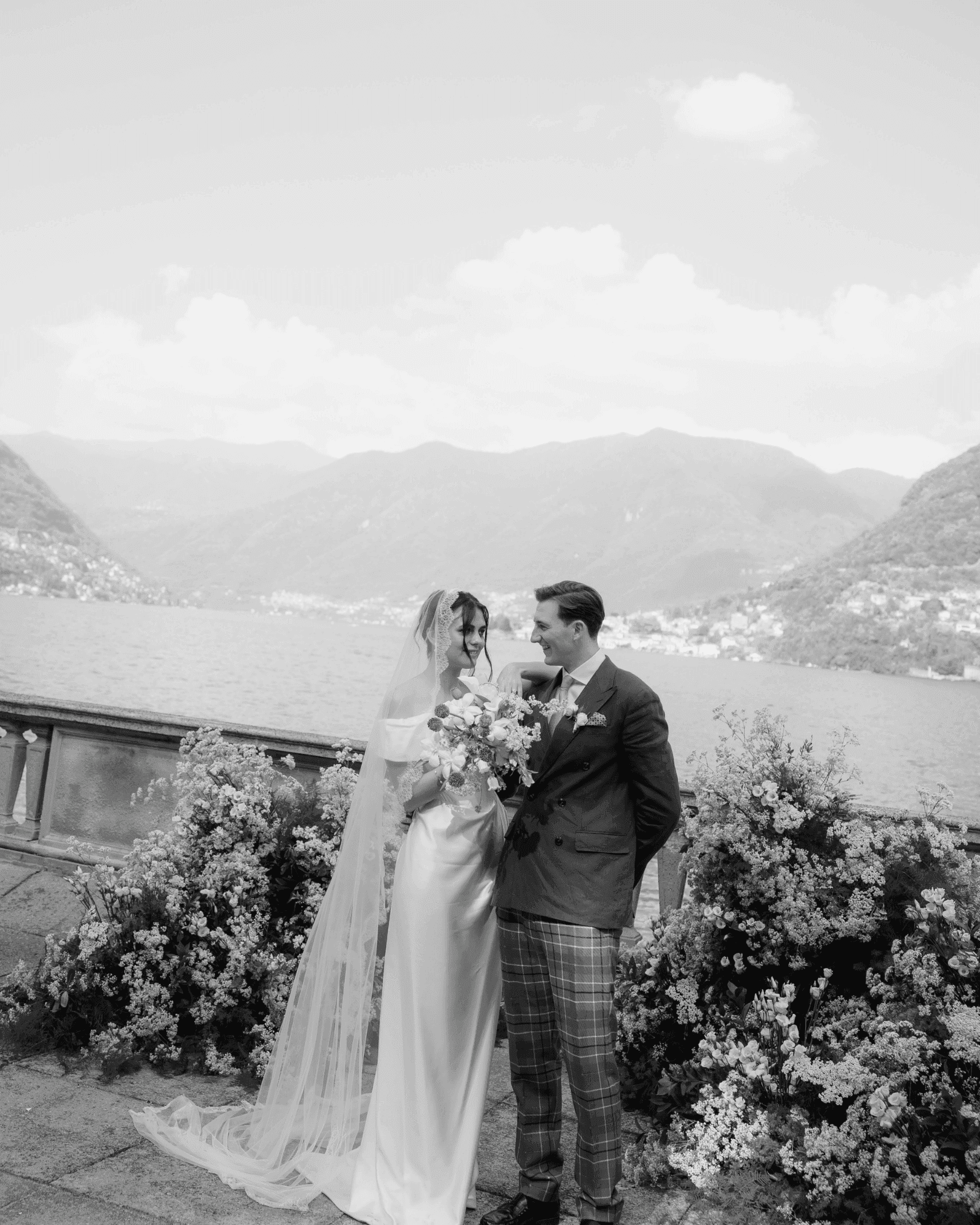 Black and white portrait of a bride and groom standing together outdoors surrounded by flowers with the lake in the background